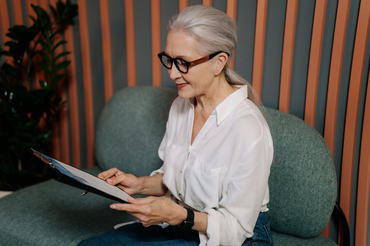 Elderly woman in glasses reviewing documents on clipboard, sitting indoors with a focused expression.